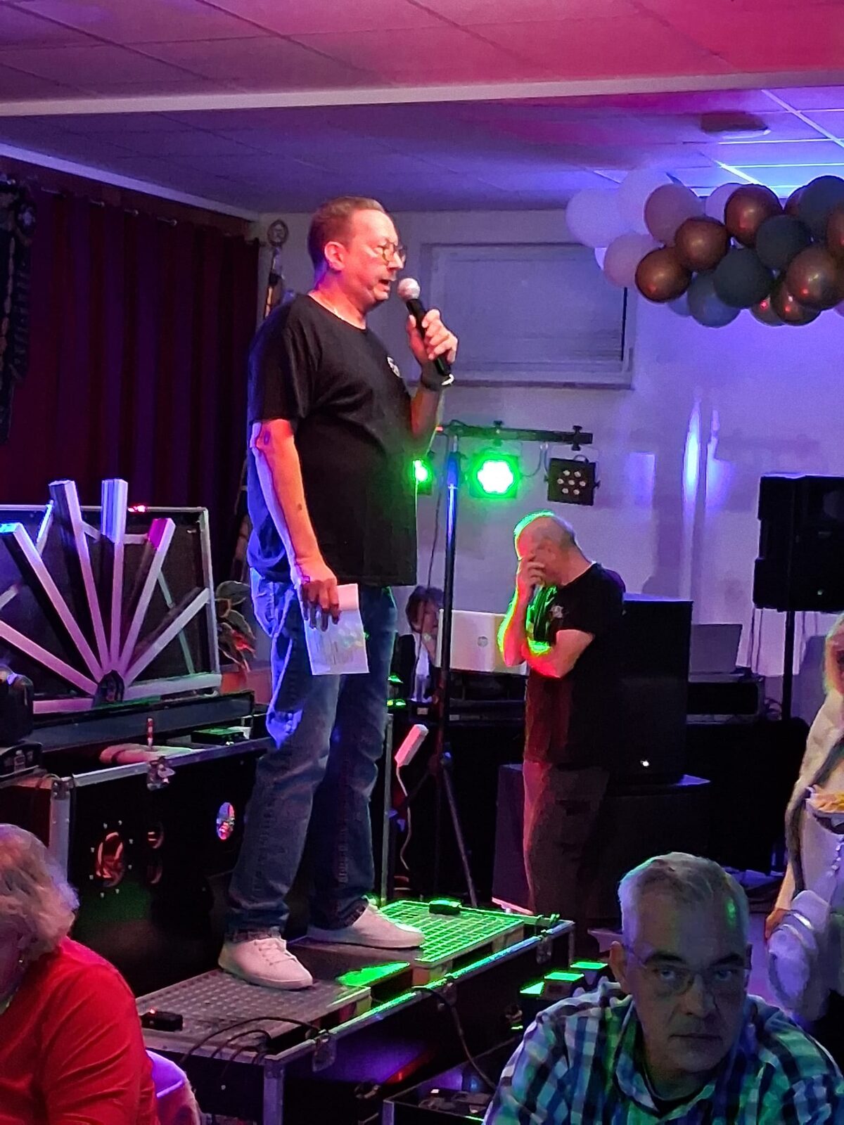 Abend im Vereinsheim: jemand am Mikrofon auf der Bühne, im Hintergrund grüne Scheinwerfer und Luftballons